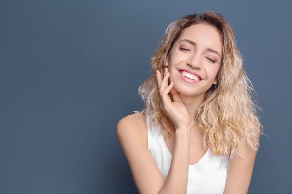 A cheerful young woman with wavy blonde hair smiles brightly while gently touching her face against a solid blue background. - Cosmetic Dentistry